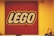 A square display of the LEGO logo composed of numerous small red, white, and yellow pieces on a red background, mounted on a yellow wall. In the foreground, there are various LEGO models and a security camera visible in the upper right corner.
