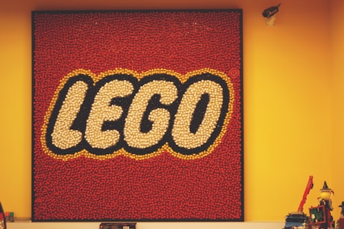 A square display of the LEGO logo composed of numerous small red, white, and yellow pieces on a red background, mounted on a yellow wall. In the foreground, there are various LEGO models and a security camera visible in the upper right corner.