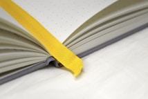An open notebook lies on a white surface, featuring a dotted page pattern. A bright yellow ribbon bookmark runs diagonally across the page, adding a pop of color against the muted tones of the paper. The notebook's spine is visible, suggesting a hardcover binding.