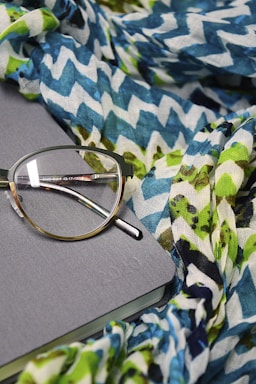 A pair of eyeglasses rests on a closed gray notebook, surrounded by a colorful, patterned fabric with zigzag designs in shades of blue, green, and white.