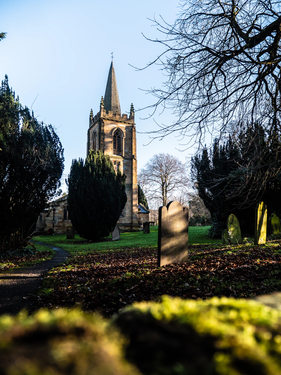 The image depicts a serene cemetery scene with a tall, pointed church steeple standing out against the clear blue sky. The church is nestled amidst lush green trees and bushes, creating a peaceful atmosphere. In front of the church, there's a small stone headstone that adds to the solemnity of the setting.

The perspective of the image is taken from ground level, looking up at the church steeple, giving it an imposing presence. The sky above is a beautiful shade of blue, providing a striking contrast to the greenery below and enhancing the overall tranquility of the scene.