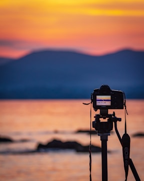 A DSLR camera mounted on a tripod captures the serene beauty of a sunset over the water, with calm waves in the foreground and silhouettes of distant hills against a colorful sky.