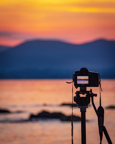 A DSLR camera mounted on a tripod captures the serene beauty of a sunset over the water, with calm waves in the foreground and silhouettes of distant hills against a colorful sky.