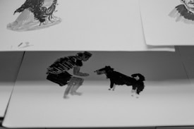 A black and white illustration on paper depicting a person crouching and extending their hand towards a dog. The artwork is simple yet expressive, focusing on the interaction between the person and the dog. The paper is slightly curled and resting on top of another sketch.
