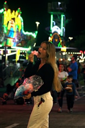A woman is enjoying a piece of cotton candy with a lively carnival scene in the background. Bright lights and colorful rides create a vibrant atmosphere. She's holding a plastic bag of cotton candy and surrounded by other people, including a child.