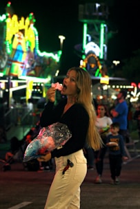 A woman is enjoying a piece of cotton candy with a lively carnival scene in the background. Bright lights and colorful rides create a vibrant atmosphere. She's holding a plastic bag of cotton candy and surrounded by other people, including a child.