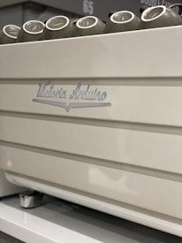 A close-up view of a white coffee machine with metallic accents, featuring cups placed on top. The machine has the brand name 'Victoria Arduino' inscribed on it.