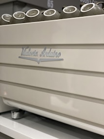 A close-up view of a white coffee machine with metallic accents, featuring cups placed on top. The machine has the brand name 'Victoria Arduino' inscribed on it.