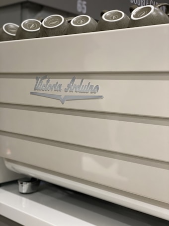 A close-up view of a white coffee machine with metallic accents, featuring cups placed on top. The machine has the brand name 'Victoria Arduino' inscribed on it.