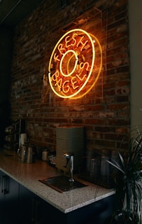 A neon sign in the shape of a bagel with the words 'Fresh Bagels' is mounted on a rustic brick wall. Below the sign is a countertop with various items including metal canisters, a stack of napkins, plastic cups, and a small plant. The overall ambiance is dimly lit, giving a cozy and inviting feel to the space.