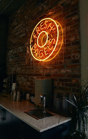 A neon sign in the shape of a bagel with the words 'Fresh Bagels' is mounted on a rustic brick wall. Below the sign is a countertop with various items including metal canisters, a stack of napkins, plastic cups, and a small plant. The overall ambiance is dimly lit, giving a cozy and inviting feel to the space.