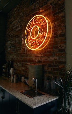 A neon sign in the shape of a bagel with the words 'Fresh Bagels' is mounted on a rustic brick wall. Below the sign is a countertop with various items including metal canisters, a stack of napkins, plastic cups, and a small plant. The overall ambiance is dimly lit, giving a cozy and inviting feel to the space.