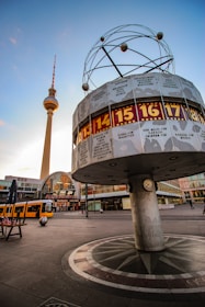 An urban scene featuring a unique world clock with a circular structure displaying various city names and times. A tall television tower with a distinct red and white antenna stands in the background. The sky has a gradient of blue and orange tones as the sun sets.