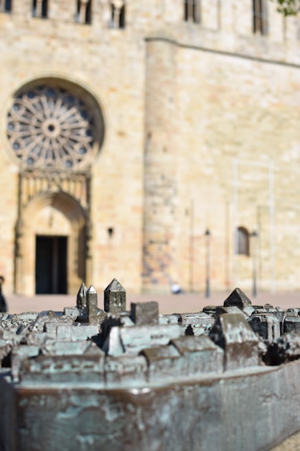 A detailed close-up of a metallic model depicting a miniature cityscape, with small buildings and structures. In the background, a large, historic building with an ornate rose window and a grand entrance made of stone is visible.
