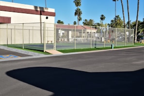 A tennis court fenced with a chain-link fence is situated next to a building with light-colored walls and a maroon trim. The area surrounding the court features palm trees and other greenery, creating a tropical atmosphere. The ground is paved, with a section marked for accessible parking. Clear skies suggest sunny weather.