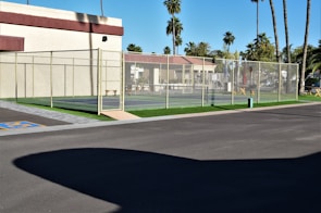 A tennis court fenced with a chain-link fence is situated next to a building with light-colored walls and a maroon trim. The area surrounding the court features palm trees and other greenery, creating a tropical atmosphere. The ground is paved, with a section marked for accessible parking. Clear skies suggest sunny weather.