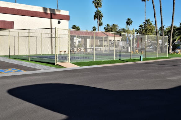 A tennis court fenced with a chain-link fence is situated next to a building with light-colored walls and a maroon trim. The area surrounding the court features palm trees and other greenery, creating a tropical atmosphere. The ground is paved, with a section marked for accessible parking. Clear skies suggest sunny weather.