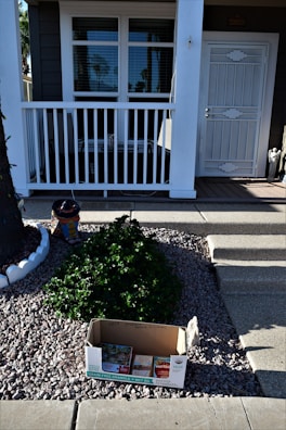 A house entrance with a covered porch and white railing. In the foreground, there's a cardboard box labeled with various food product logos placed on a gravel pathway. Behind the box is a small, manicured shrub and a decorative pot shaped like a penguin. The porch has closed windows with blinds and a white, paneled front door.