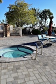 A whirlpool with blue handrails is surrounded by a paved patio area. Nearby, there are several outdoor chairs around a table with an umbrella. The area is shaded by various trees, including a large tree with dense foliage and several tall palm trees in the background. Bright sunlight casts shadows on the ground.