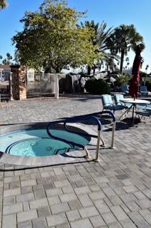 A whirlpool with blue handrails is surrounded by a paved patio area. Nearby, there are several outdoor chairs around a table with an umbrella. The area is shaded by various trees, including a large tree with dense foliage and several tall palm trees in the background. Bright sunlight casts shadows on the ground.
