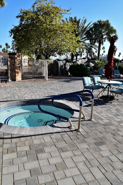 A whirlpool with blue handrails is surrounded by a paved patio area. Nearby, there are several outdoor chairs around a table with an umbrella. The area is shaded by various trees, including a large tree with dense foliage and several tall palm trees in the background. Bright sunlight casts shadows on the ground.