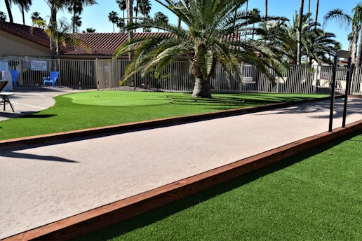 A landscaped area with a palm tree surrounded by artificial grass, a pathway with wood edging, and a few blue chairs in the background. A putting green with small flags is situated next to a fence with a red-roofed building behind it.