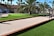 A landscaped area with a palm tree surrounded by artificial grass, a pathway with wood edging, and a few blue chairs in the background. A putting green with small flags is situated next to a fence with a red-roofed building behind it.