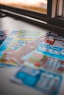 A selection of colorful lottery scratch-off tickets placed on a flat surface near a window. The image has a shallow depth of field, focusing on the tickets while blurring the background. The tickets display various games and prize amounts.