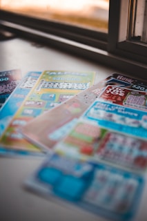 A selection of colorful lottery scratch-off tickets placed on a flat surface near a window. The image has a shallow depth of field, focusing on the tickets while blurring the background. The tickets display various games and prize amounts.