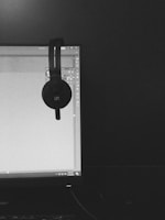 A black and white photograph features a computer monitor displaying a software interface, likely related to graphic design. A pair of over-ear headphones is hanging from the top corner of the monitor. The room appears dimly lit, and the overall setup conveys a workspace.