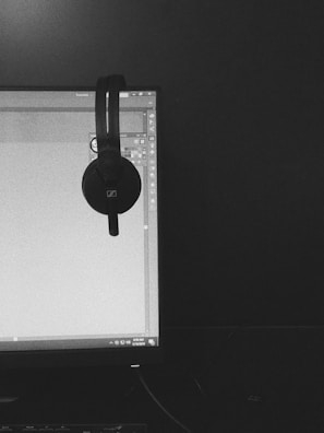 A black and white photograph features a computer monitor displaying a software interface, likely related to graphic design. A pair of over-ear headphones is hanging from the top corner of the monitor. The room appears dimly lit, and the overall setup conveys a workspace.