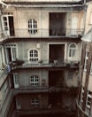 A multi-story residential building with a central courtyard surrounded by several floors of narrow balconies. The walls are gray with large rectangular windows, and there are various plants and bicycles on the balconies. The structure has arched doorways and windows lined with metal railings.