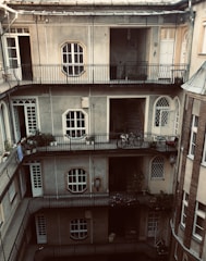 A multi-story residential building with a central courtyard surrounded by several floors of narrow balconies. The walls are gray with large rectangular windows, and there are various plants and bicycles on the balconies. The structure has arched doorways and windows lined with metal railings.