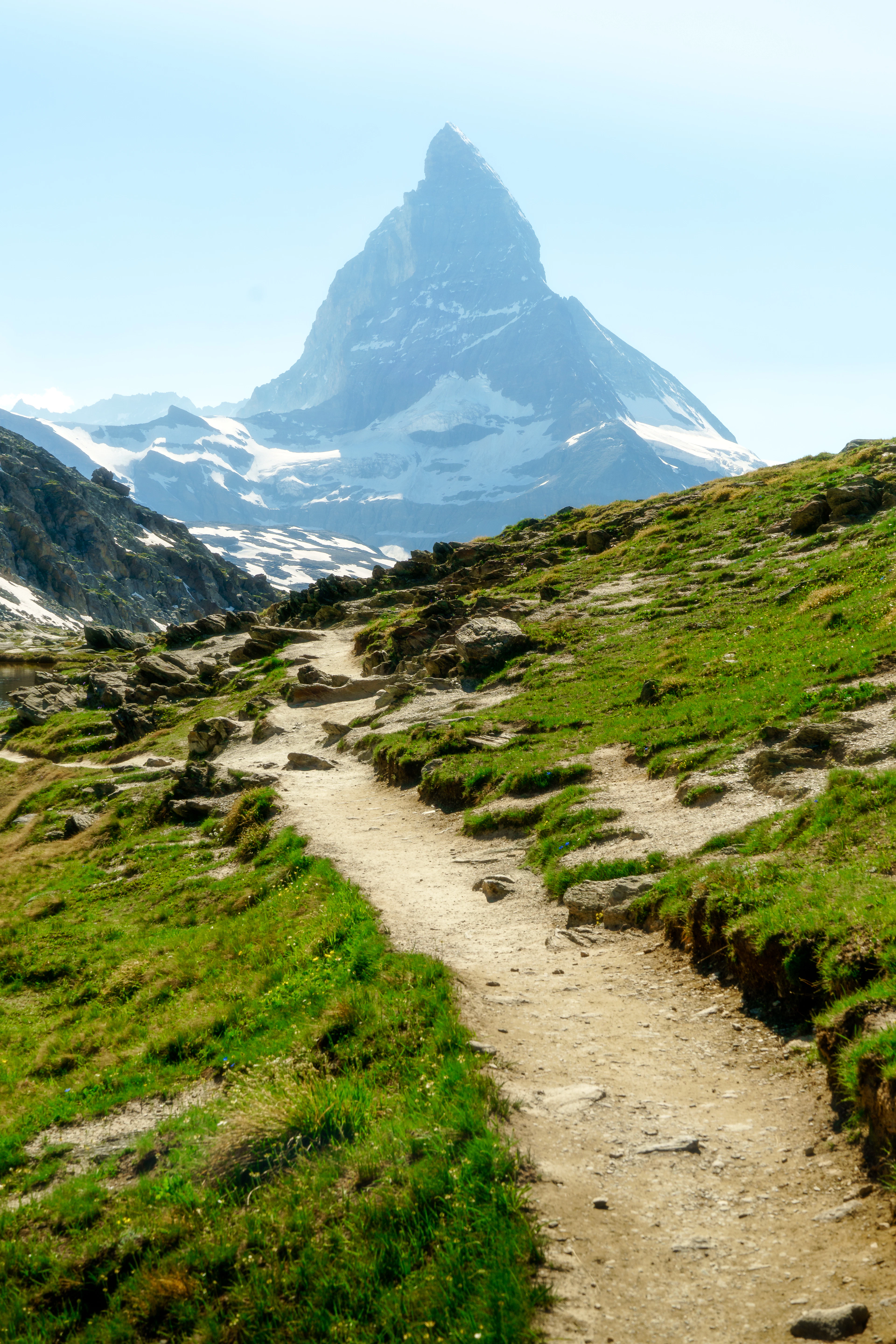 A grassy hiking trail leads straight toward the Matterhorn's peak under a bright sky.
