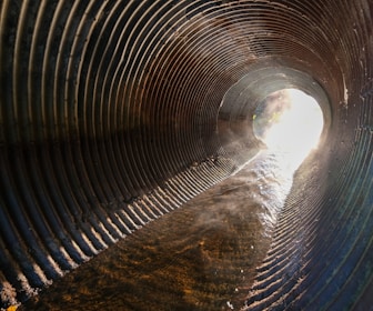 A tunnel with corrugated metal walls and water flowing through it. Sunlight is streaming in from the opening, casting a glow and creating a misty effect. Ripples in the water are visible, along with reflections on the wet metal surface.