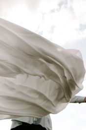 A flowing, translucent piece of fabric billows gracefully in the wind against a partially cloudy sky. The fabric appears to be light and airy, capturing the sunlight as it moves.