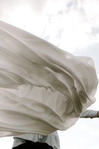 A flowing, translucent piece of fabric billows gracefully in the wind against a partially cloudy sky. The fabric appears to be light and airy, capturing the sunlight as it moves.