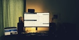 A dimly lit workspace featuring a large computer monitor displaying multiple charts and diagrams. The monitor is flanked by speakers and a small figurine on the right. There is also a tablet on the left with a visible webpage. The lighting is soft and the room appears calm with minimal clutter.