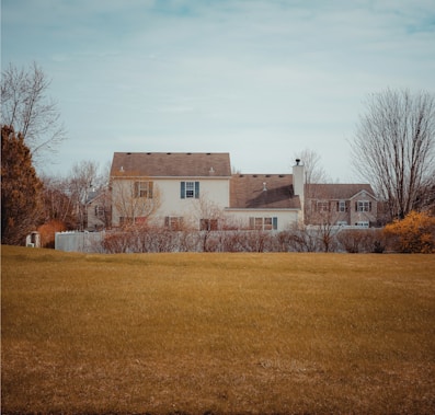A suburban house is seen in the background with a large, open lawn in the foreground. The house has a two-story structure with beige siding and a brown roof. Surrounding the house are several bare trees and bushes, suggesting it might be late autumn or early winter. The sky is overcast with a slight blue tint.