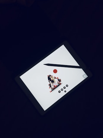 A tablet lies flat on a dark surface, displaying an image of a woman adorned with flowers. A stylus pen is placed horizontally across the screen. The tablet screen is mainly white with minimalistic design elements, including a red circle and several black icons at the bottom.