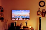 A living room setting is stylishly decorated with a large wall-mounted TV displaying an image of boats moored in a calm harbor under a colorful sky. Below the TV, a table holds a decorative Buddha head, vases with flowers, and a Himalayan salt lamp. Shelves on both sides feature books and small decorative items. The warm lighting creates a cozy atmosphere.