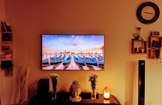 A living room setting is stylishly decorated with a large wall-mounted TV displaying an image of boats moored in a calm harbor under a colorful sky. Below the TV, a table holds a decorative Buddha head, vases with flowers, and a Himalayan salt lamp. Shelves on both sides feature books and small decorative items. The warm lighting creates a cozy atmosphere.
