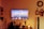 A living room setting is stylishly decorated with a large wall-mounted TV displaying an image of boats moored in a calm harbor under a colorful sky. Below the TV, a table holds a decorative Buddha head, vases with flowers, and a Himalayan salt lamp. Shelves on both sides feature books and small decorative items. The warm lighting creates a cozy atmosphere.