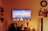 A living room setting is stylishly decorated with a large wall-mounted TV displaying an image of boats moored in a calm harbor under a colorful sky. Below the TV, a table holds a decorative Buddha head, vases with flowers, and a Himalayan salt lamp. Shelves on both sides feature books and small decorative items. The warm lighting creates a cozy atmosphere.