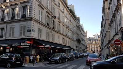 A Parisian street scene with classic Haussmann-style buildings lining both sides of a narrow, one-way street. The buildings have many windows with shutters and black wrought-iron balconies. On the left corner, a bar and restaurant named Le Cardinal features outdoor seating with people dining. Several parked cars are visible along the street, and there is a clear blue sky overhead.