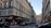 A Parisian street scene with classic Haussmann-style buildings lining both sides of a narrow, one-way street. The buildings have many windows with shutters and black wrought-iron balconies. On the left corner, a bar and restaurant named Le Cardinal features outdoor seating with people dining. Several parked cars are visible along the street, and there is a clear blue sky overhead.