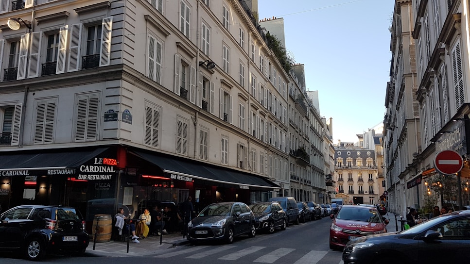 A Parisian street scene with classic Haussmann-style buildings lining both sides of a narrow, one-way street. The buildings have many windows with shutters and black wrought-iron balconies. On the left corner, a bar and restaurant named Le Cardinal features outdoor seating with people dining. Several parked cars are visible along the street, and there is a clear blue sky overhead.