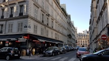 A Parisian street scene with classic Haussmann-style buildings lining both sides of a narrow, one-way street. The buildings have many windows with shutters and black wrought-iron balconies. On the left corner, a bar and restaurant named Le Cardinal features outdoor seating with people dining. Several parked cars are visible along the street, and there is a clear blue sky overhead.