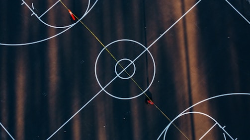 An aerial view of a basketball court with distinct white markings and two red cones positioned near the center circle. The court is surrounded by long shadows, creating a pattern across the surface.