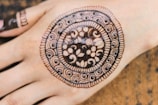 An intricately designed mehndi pattern adorns the back of a hand, featuring concentric circles with floral and spiral motifs, bordered by a detailed lace-like edge.